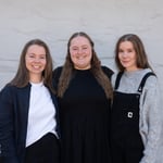 Tre unge personer står foran en grå vegg. De smiler alle mot kamera