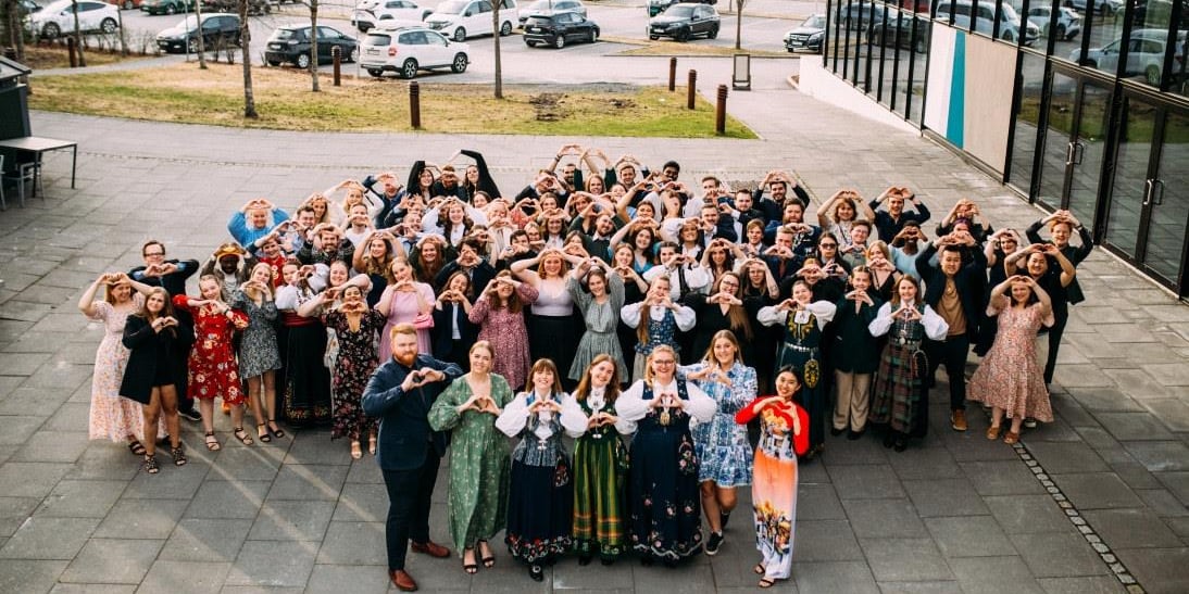 Bilde fra Pedagogstudentenes landsmøte 2023. Alle deltakerne, rundt 100 stk, står samlet på et åpent område utenfor Sundvolden Hotell. De ser opp mot fotografen, som står på en høyde og tar bilde av menneskene. De holder hendene opp og lager et hjerte. Menneskene er pyntet i dress, kjoler og bunader. Foto.