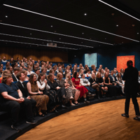 En person står på en scene foran et publikum i et auditorium med amfi. Publikum sitter i flere rekker, og rommet er dempet belyst med spotlights som lyser opp scenen og deler av salen. Tre fargerike, abstrakte malerier henger på veggen bak publikum.
