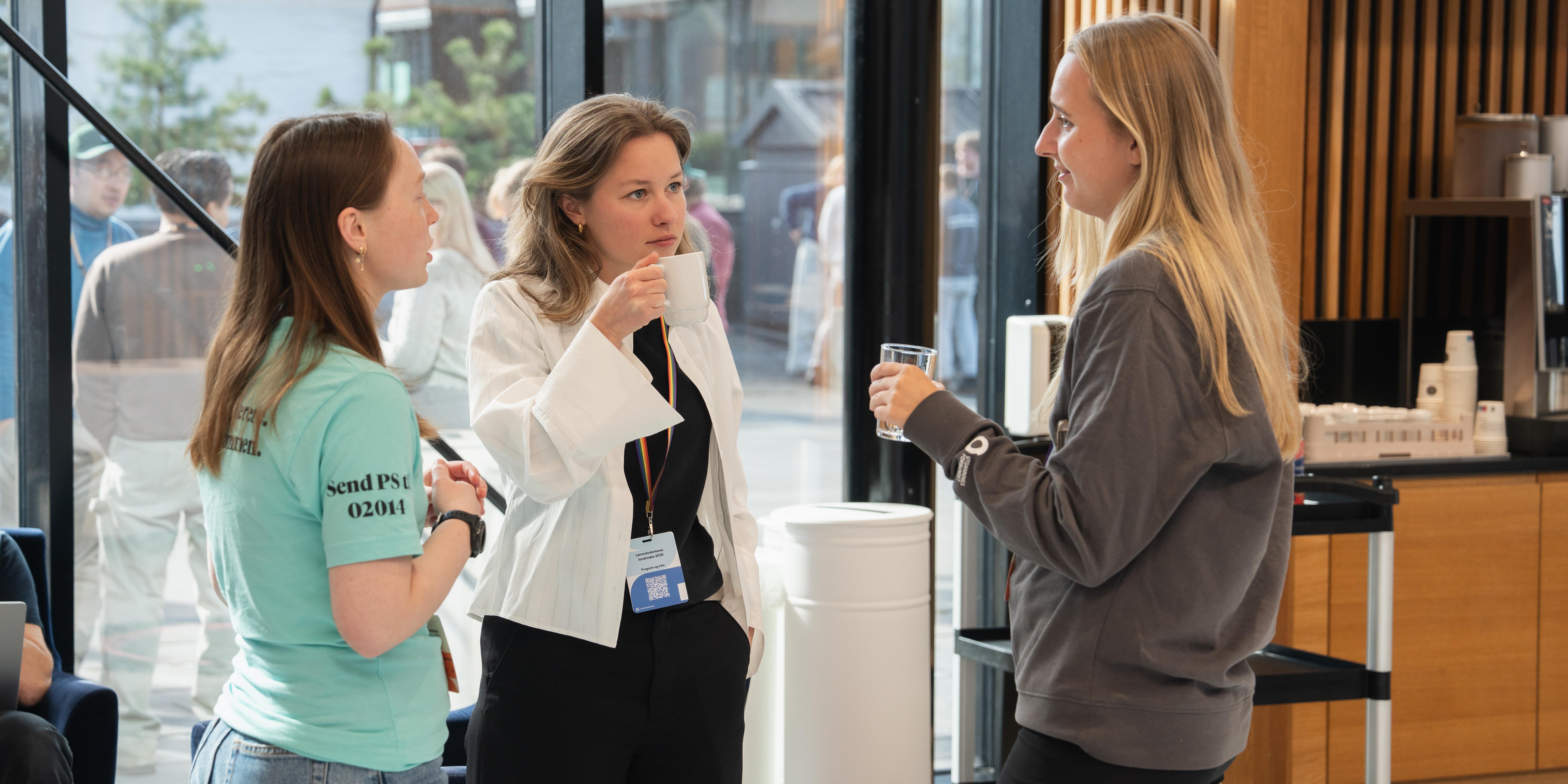 Tre kvinner snakker sammen i en pause under et arrangement. To holder drikke, og de står ved en kaffestasjon i et lyst rom med store vinduer.
