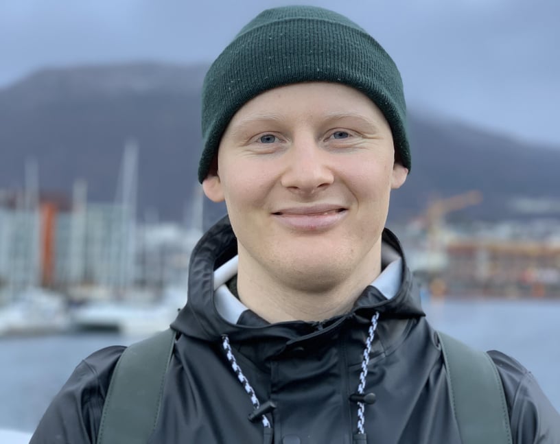 Gutt står foran kamera og smiler. Han har på seg lue og regnjakke. I bakgrunnen kan du se fjell og båter.