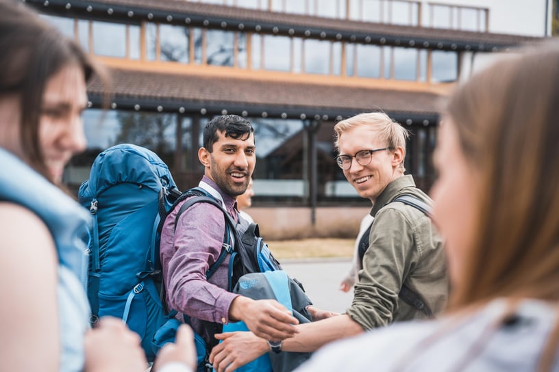 Bilde av fire personer, hvor to av de er i fokus i bakgrunn. Personene snakker med hverandre og smiler. De har på seg sekk og ser ut til å være klare for å reise. 