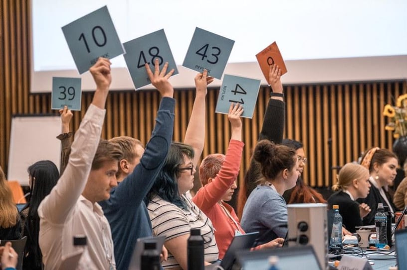 Bilde av delegater på landsmøte, som strekker stemmeskilt i været. 