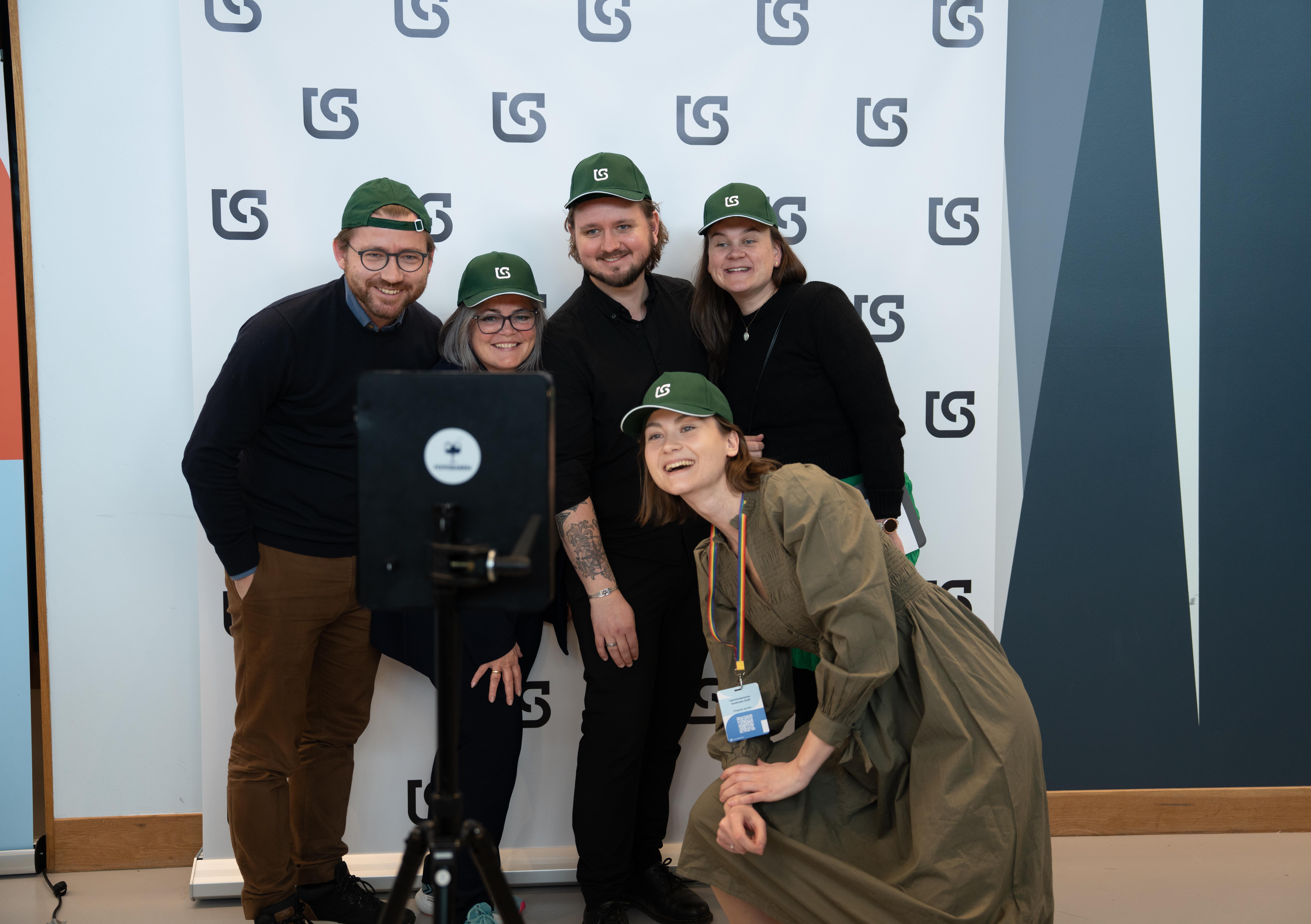 Fem personer står og smiler inn i et kamera. Alle har på seg like, grønne capser.