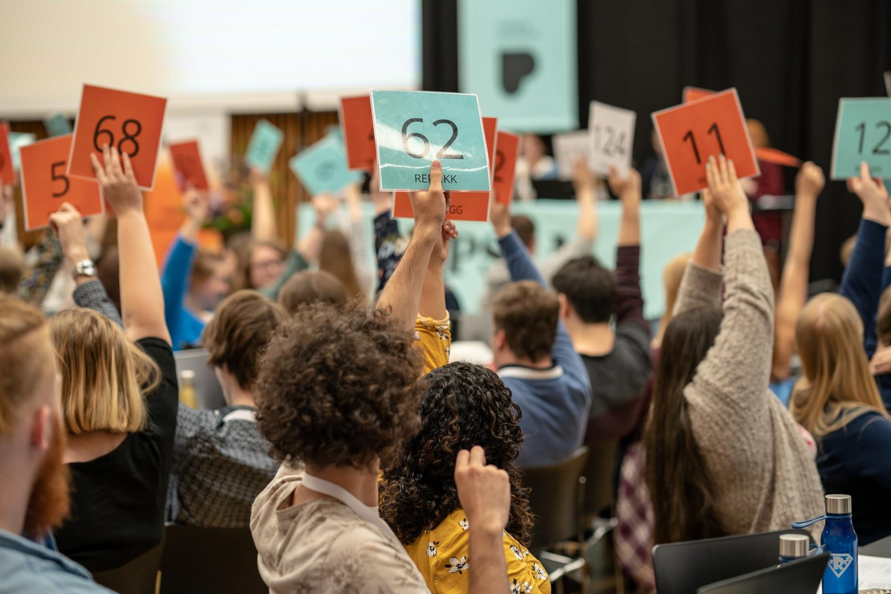 Mange mennesker som sitter i en sal med ryggen mot kamera. De fleste holder opp stemmeskilt. Bildet er tatt under landsmøtet til Pedagogstudentene i 2019. Foto: Chris-Håvard Berge/Oclin