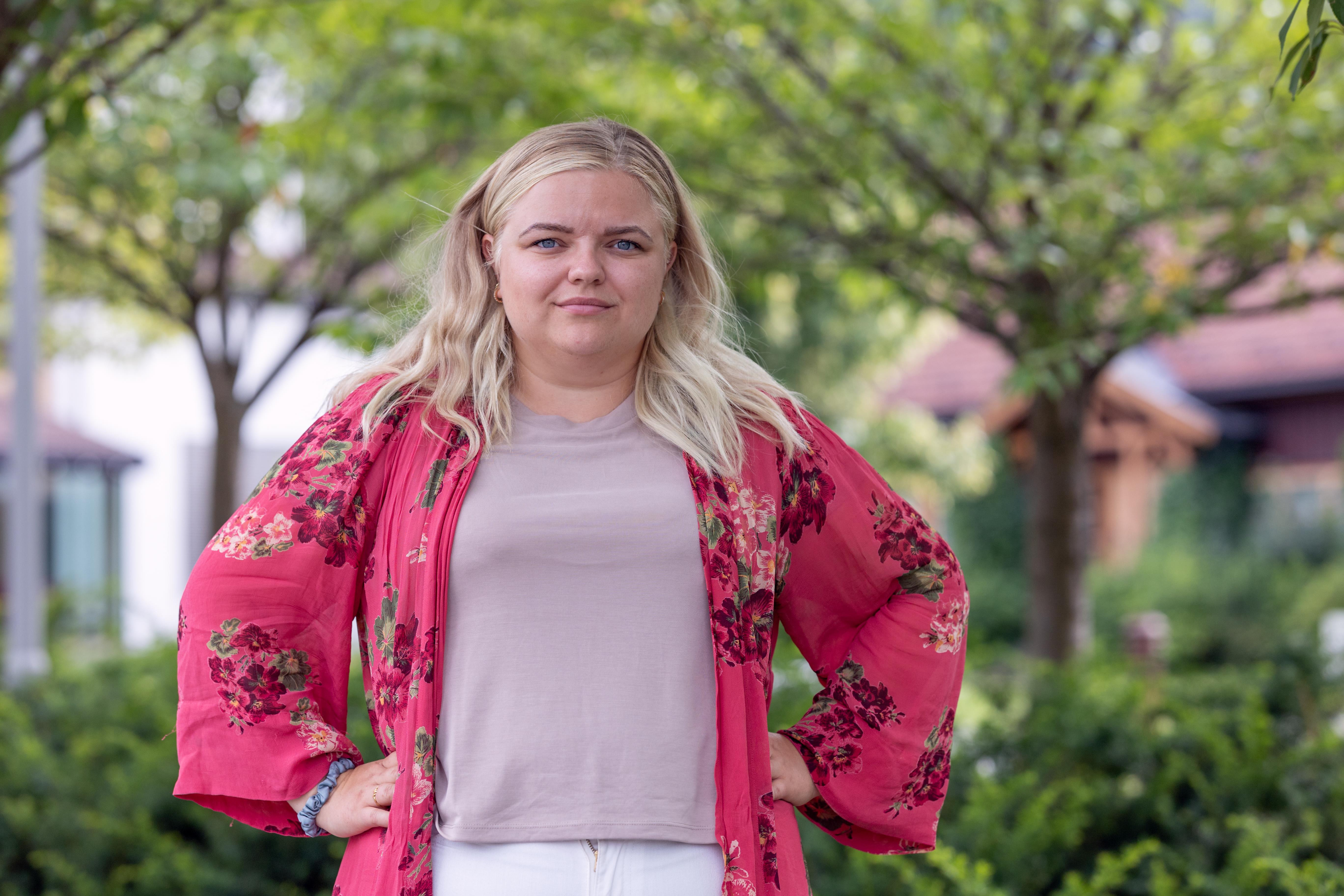 Blond kvinne står med hendene på hoftene og ser inn i kamera. I bakgrunnen er det grønne trær og kvinnen har på seg rosa klær. Foto. 